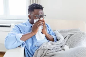 Ill African young woman covered with blanket blowing running nose got fever caught cold sneezing in tissue sit on sofa, sick allergic black girl having allergy symptoms coughing at home, flu concept. 