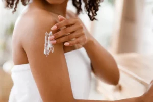 Skin Care Products Concept. Black woman applying moisturising lotion on body after shower, standing wrapped in towel, cropped image. 