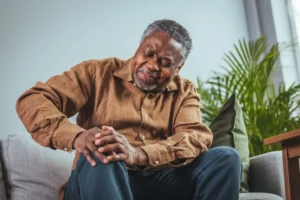 Photo of mature, elderly man sitting on a sofa in the living room at home and touching his knee by the pain during the day. Mature man massaging his painful knee. 