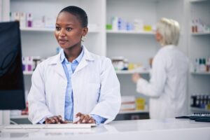 Pharmacist, typing and black woman on computer in pharmacy, drugstore or shop. Wellness, medical professional and African doctor on internet for telehealth email, healthcare and research for medicine. 