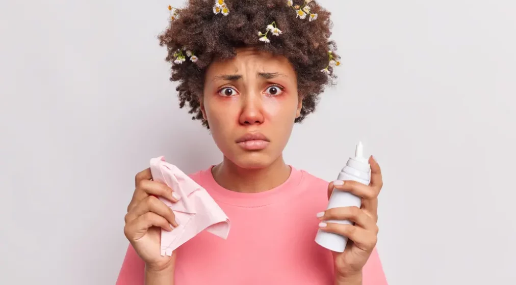 Disappointed young Afro American woman uses nasal aerosol suffers from allergic rhinitis has red swollen eyes looks unhappily at camera dressed in casual t shirt isolated over white background.