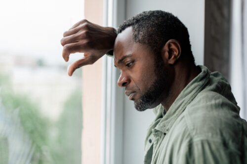 Unhappy frustrated adult African guy in casual suffering from depression and bad news near window in home interior. Health problems, stress from self-isolation during covid-19 quarantine.