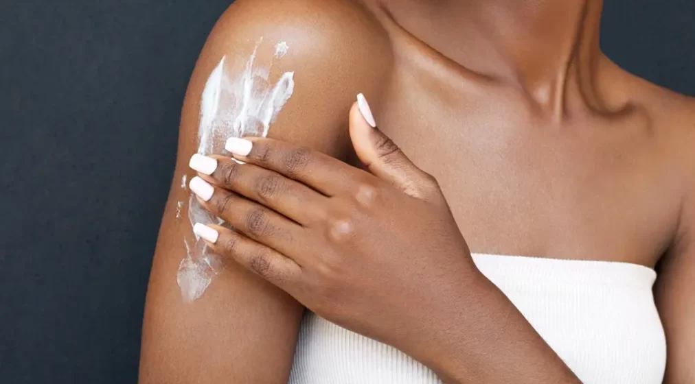 Body care. Hydration. Dry skin. African American young woman is applying skincare lotion on her arms and smiling. Moisturizing and rejuvenation beauty product. Spa treatment, scrub, peeling.