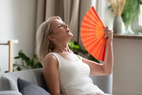 Stressed annoyed old senior woman using waving fan suffer from overheating, summer heat health hormone problem, no air conditioner at home sit on sofa feel exhaustion dehydration heatstroke concept.