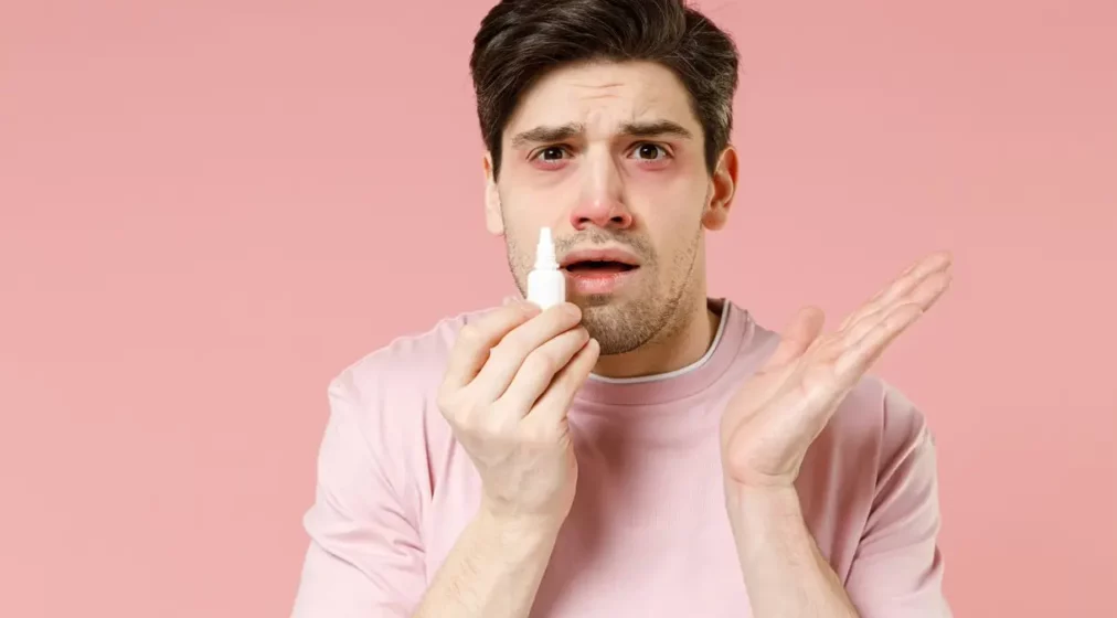 Sick unhealthy ill allergic man has red watery eyes runny stuffy sore nose suffer from allergy symptoms hay fever hold bottle uses nasal drops isolated on pastel pink colour background studio.