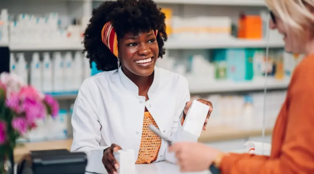 Portrait of a smiling African woman pharmacist working in a pharmacy while holding medication in her hands and leaning on a counter with a cash register.