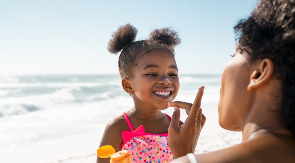 Young mother applying protective sunscreen on daughter nose at beach with copy space. Black woman hand putting sun lotion on female child face. African American cute little girl with sunblock cream.