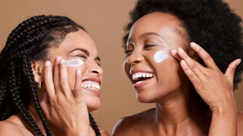 Black woman, cream and skincare with friends support, love and beauty together in studio for cosmetics, makeup cleaning and facial. Excited, diversity model with sunscreen or essential oil on face.