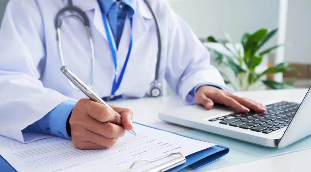 hands-unrecognizable-female-doctor-writing-form-typing-laptop-keyboard